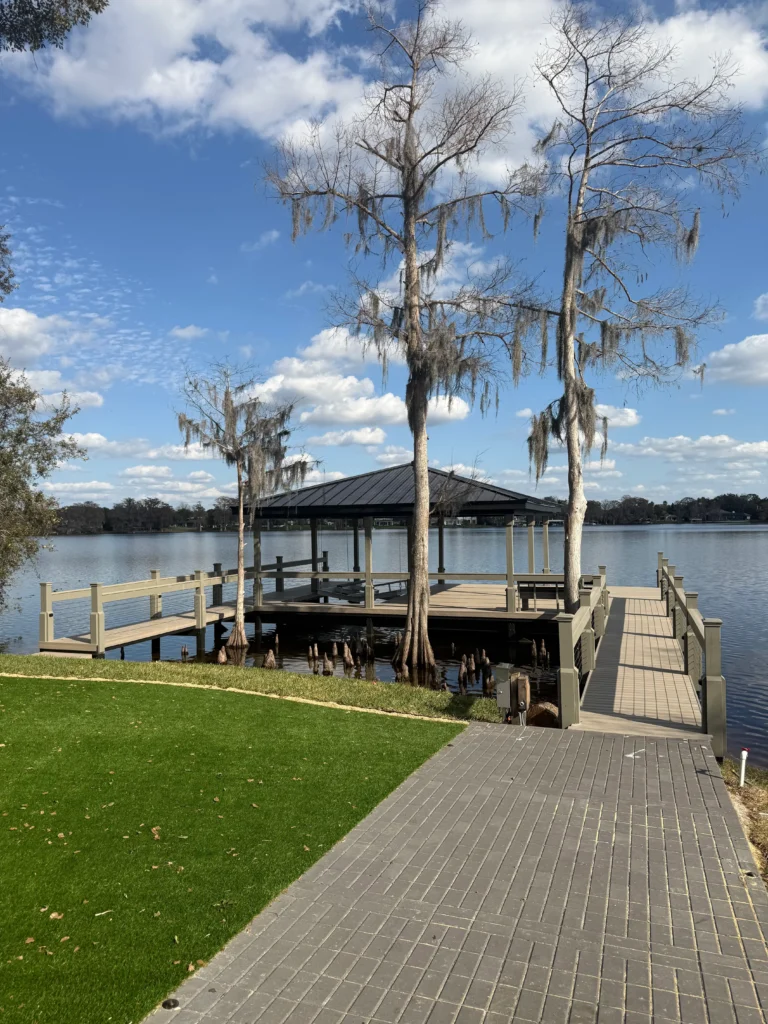 A dock extending from a brick pathway, flanked by trees and calm lake water.