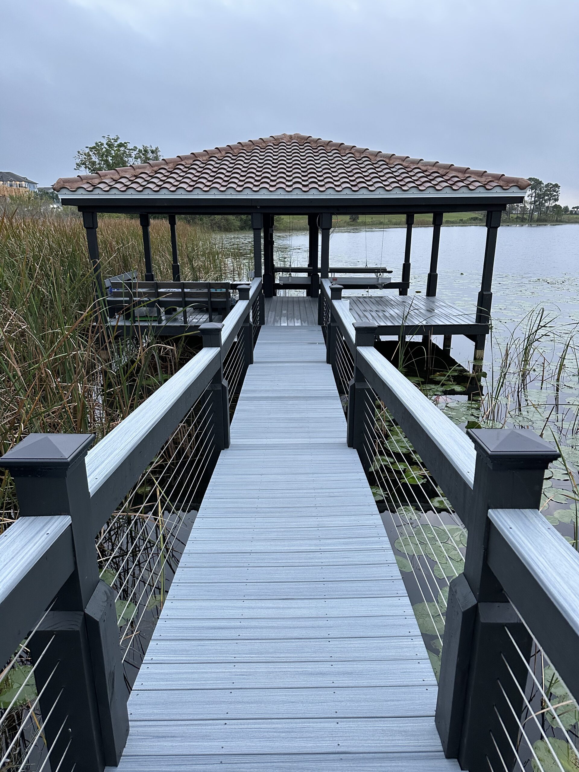 Gray boardwalk dock with terracotta-roofed boathouse extending into marshy lake.